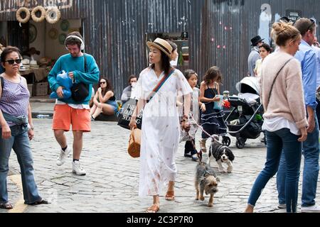 London, Großbritannien - 17. Juli 2019, Columbia Road Flower Market. Eine Frau in einem langen weißen Kleid, ein Strohhut mit zwei Hunden an der Leine geht durch die Krähe Stockfoto