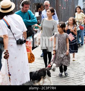 London, Großbritannien - 17. Juli 2019, Columbia Road Flower Market. Eine Frau in einem langen weißen Kleid, ein Strohhut mit zwei Hunden an der Leine geht durch die Krähe Stockfoto