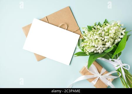 Leere Grußkarte mit Maiglöckchen Blumenstrauß und Geschenkbox auf blauem Hintergrund. Mock up. Flat lay. Draufsicht. Hochzeitseinladung Stockfoto