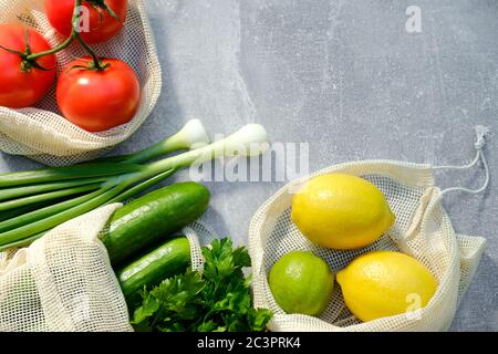Mesh produzieren Beutel mit frischem Gemüse und Obst auf Stein Beton Tisch. Draufsicht Tomaten, Gurken, Zwiebeln, Petersilie, Zitrone und Limette. Gesundheitsorgan Stockfoto