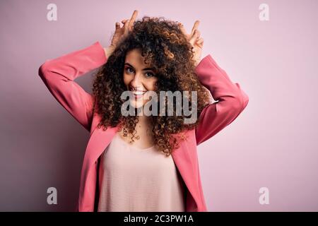 Junge schöne Geschäftsfrau mit lockigem Haar und Piercing tragen elegante Jacke posiert lustig und verrückt mit Fingern auf dem Kopf als Hase Ohren, lächelnd c Stockfoto