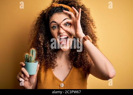 Junge schöne Frau mit lockigen Haaren und stechenden Haltestopf mit Kakteenpflanze mit fröhlichem Gesicht lächelnd, dabei ok Schild mit Hand auf Auge durch Stockfoto