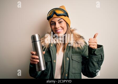 Junge schöne Brünette Skifahrer Frau trägt Skibrille trinken Thermo mit Kaffee glücklich mit großen Lächeln tun ok Zeichen, Daumen hoch mit Fingern, excelle Stockfoto