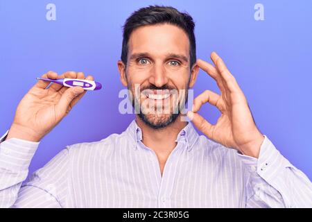 Junge gut aussehende Mann Controlling Temperatur halten Thermometer über lila Hintergrund tun ok Zeichen mit Fingern, lächelnd freundlich gestikulieren ausgezeichnet Stockfoto