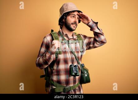Junger Wanderer mit lockigem Haar und Bart, der Rucksack und Wasserkantine trägt, ist sehr glücklich und lächelnd und schaut mit der Hand über dem Kopf weit weg. Such Stockfoto