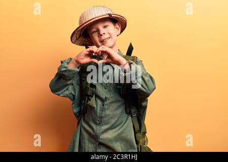 Cute blonde Kind trägt Explorer Hut und Rucksack lächelnd in der Liebe tun Herz Symbol Form mit Händen. Romantisches Konzept. Stockfoto