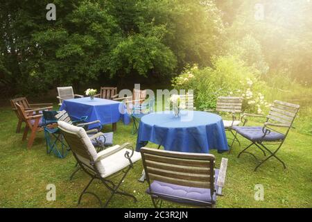 Richten Sie sich für eine private Gartenparty mit Tischen, blauen Tischdecken und vielen verschiedenen Stühlen in einem ländlichen Hinterhof in goldenen späten Nachmittag Sonnenlicht, selec Stockfoto
