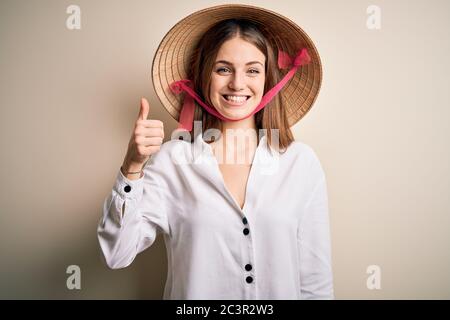 Junge schöne Rothaarige Frau trägt asiatische traditionelle konische Hut über weißem Hintergrund tun glücklich Daumen nach oben Geste mit der Hand. Genehmigung des Expressio Stockfoto