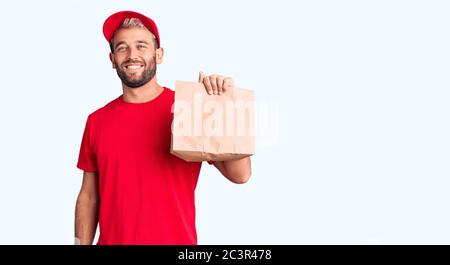 Junge gut aussehende blonde Mann hält Lieferung Papiertasche suchen positiv und glücklich stehend und lächelnd mit einem selbstbewussten Lächeln zeigt Zähne Stockfoto