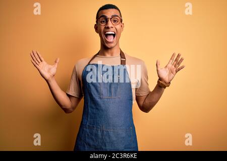 Junge gutaussehende afroamerikanische Ladenbesitzer Mann trägt Schürze über gelben Hintergrund feiert verrückt und erstaunt für den Erfolg mit den Armen angehoben und offen Stockfoto