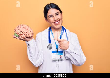 Junge schöne Psychiaterin Frau trägt Stethoskop hält Gehirn über gelben Hintergrund lächelnd glücklich zeigt mit Hand und Finger Stockfoto