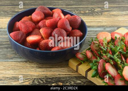 Frische, rohe Erdbeeren in einer großen blauen Schüssel und grüne, belaubte Erdbeerspitzen auf einem Holzschneidebrett Stockfoto