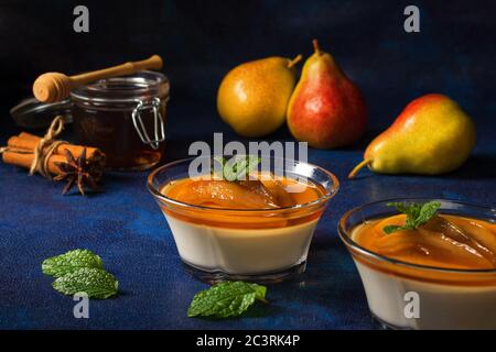 Panna Cotta Italienische traditionelle Dessert mit Birnen pochiert in Rum und Honig Sirup. Dunkelblauer Hintergrund Tisch, Gläser Rum, Glas Honig, Birnen und Stockfoto