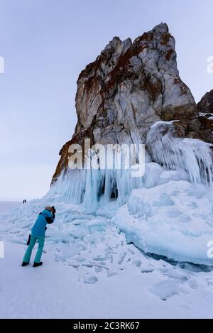 Baikalsee, Russland - 12. März 2020: Frau in blauer Jacke fotografiert eisige Felsen mit Eiszapfen am Wintersee Baikalsee Stockfoto