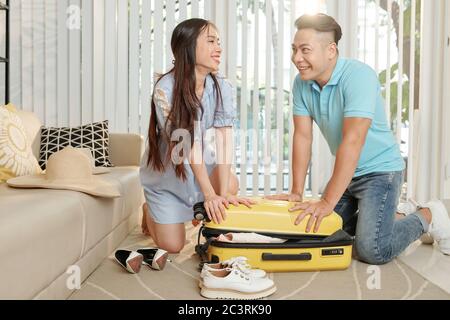 Lächelnd asiatische Freund und Freundin schieben Koffer Abdeckung, wenn kämpfen, um alle Kleidung und Schuhe sie mit ihnen nehmen wollen passen Stockfoto