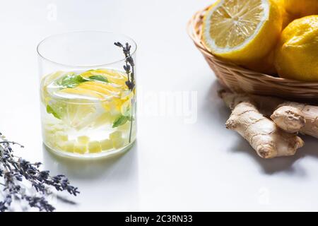 Frische Ingwerlimonade im Glas mit Minze und Lavendelzweig in der Nähe von Zitronenkorb und Ingwerwurzel auf weißem Hintergrund Stockfoto