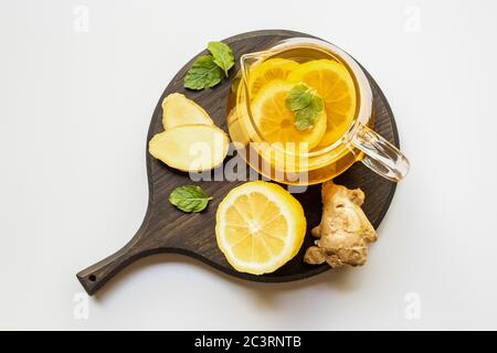 Blick von oben auf heißen Tee in Teekanne serviert auf Holzbrett mit Ingwerwurzel, Zitrone und Minze auf weißem Hintergrund Stockfoto