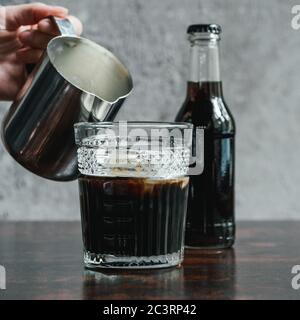 Beschnittene Ansicht der Frau, die Milch in kaltem Kaffee in Glas neben der Flasche auf Holztisch gießt Stockfoto