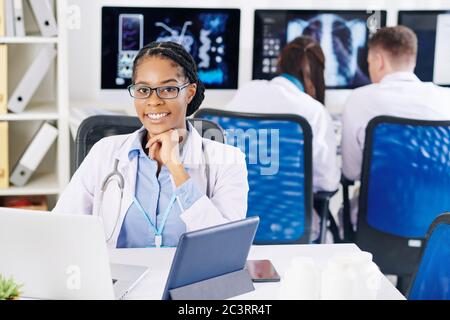 Porträt eines jungen lächelnden Arztes in einer Brille, die am Schreibtisch sitzt und am Laptop arbeitet Stockfoto