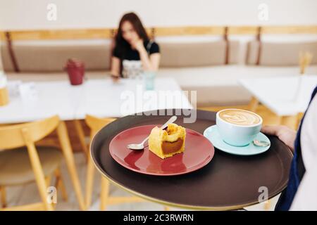 Kellner bringt Tablett mit einer Tasse Kaffee und süßem Dessert zu weiblichen Kunden, selektive Konzentration Stockfoto