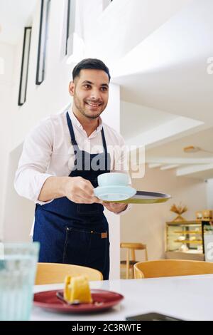 Gutaussehende lächelnde Kellner mit einem schönen Lächeln bringt Tasse Cappuccino und ein Stück Kuchen für den Kunden Stockfoto