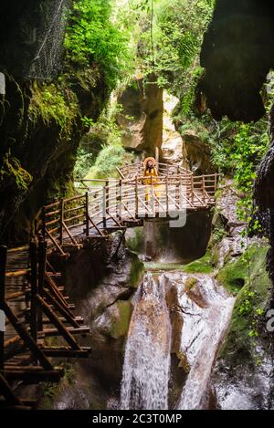 Junge wundervolle Frau in gelber Sundress in der Nähe des Wasserfalls, die auf einem Holzfußpfad zu Höhlen führt Stockfoto