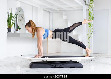 Sportlich fit junge Frau tut eine fit Surf-Plank Kick back Yoga Pose auf einem Brett in einer Seitenansicht in einem High-Key-Fitnessstudio mit frischen grünen Pflanzen in einem Gesundheits-und Stockfoto