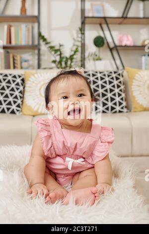 Liebenswert glücklich kleine vietnamesische Baby-Mädchen in hellrosa Kleid sitzt auf pelzigen Teppich zu Hause Stockfoto