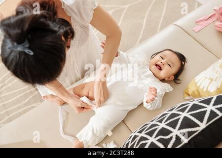 Junge Mutter Dressing ihre liebenswert lächelnd Baby Mädchen in weißen Bodysuit mit Spitze Stockfoto