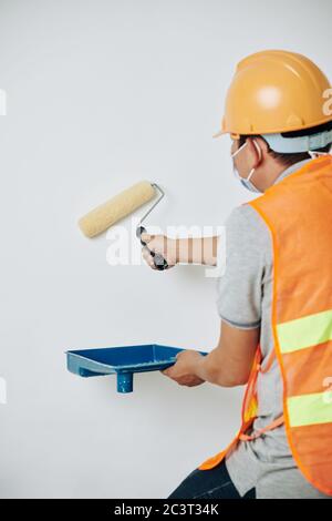 Arbeiter in Hardhat und Schutzmaske mit Kunststoff-Tablett und flauschige Rolle beim Lackieren Wände weiß Stockfoto