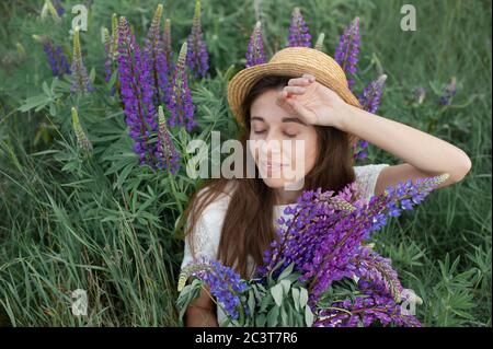 Schöne romantische Frau mit Blumenstrauß Lupinen lächelt freudig in weißem Kleid und Hut sitzt im Feld der lila Lupine Blumen. Weicher selektiver Fokus Stockfoto