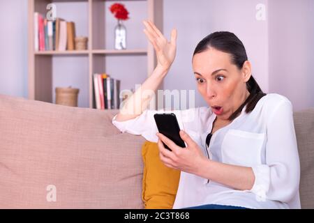 Schöne junge Brünette Frau schockiert erhält Nachrichten durch das mobile sitzen auf dem Sofa zu Hause. Sie trägt ein weißes Hemd und einen Pferdeschwanz. Stockfoto