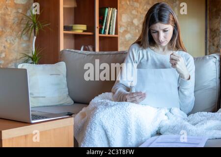 Serious junge Brünette Frau auf ein Papier sitzen auf dem Sofa zu Hause mit einer Decke Stockfoto