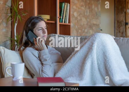 Junge ziemlich ernst Brünette Frau im Gespräch auf dem Smartphone sitzen auf dem Sofa zu Hause mit einer Decke bedeckt Blick zum Horizont. Stockfoto