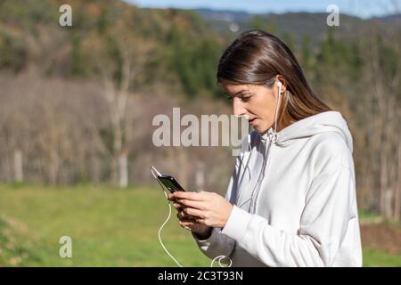 Schöne überrascht Brünette Frau liest Nachrichten auf dem Smartphone in einem Park an einem sonnigen Tag. Sie trägt einen weißen Pullover und hat lange Haare. Stockfoto