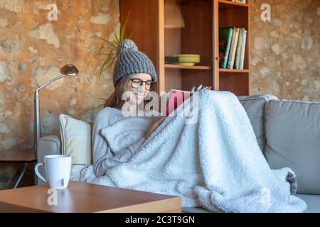 Hübsche junge Frau, die lächelt und zu Hause auf dem Sofa sitzt und ein Buch liest. Sie trägt einen Wollhut und eine Brille. Horizontales Bild Stockfoto