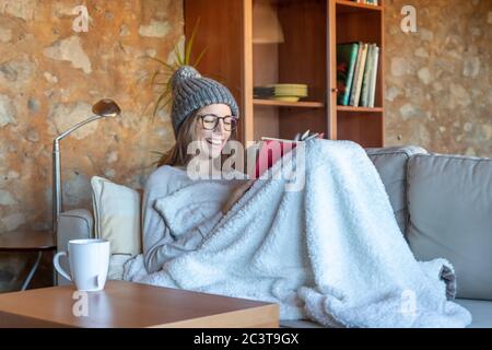 Hübsche junge Frau, die lachend auf dem Sofa zu Hause sitzt und ein Buch liest. Sie trägt einen Wollhut und eine Brille. Horizontales Bild Stockfoto