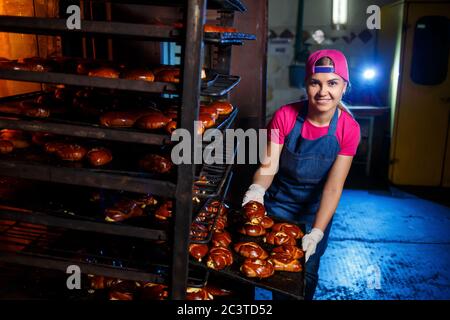 Mädchen Bäcker hält ein Tablett mit heißem Gebäck in der Bäckerei. Herstellung von Backwaren. Frische knusprige Gebäckteigablage Stockfoto