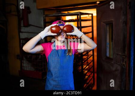 Eine professionelle Bäckerin hält frische Brötchen in den Händen und hört ihren Geruch in der Bäckerei. Sie trägt ein Jeanskittel und eine Mütze. Herstellung von BA Stockfoto