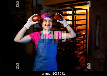 Eine professionelle Bäckerin hält frische Brötchen in den Händen und hört ihren Geruch in der Bäckerei. Sie trägt ein Jeanskittel und eine Mütze. Herstellung von BA Stockfoto
