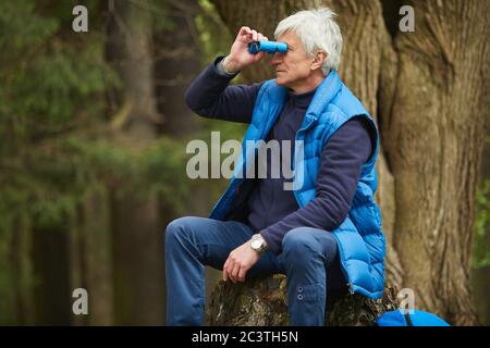 Portrait eines aktiven älteren Mannes, der im Fernglas beim Sitzen am Baum während der Wanderung im Herbstwald, Kopierraum schaut Stockfoto