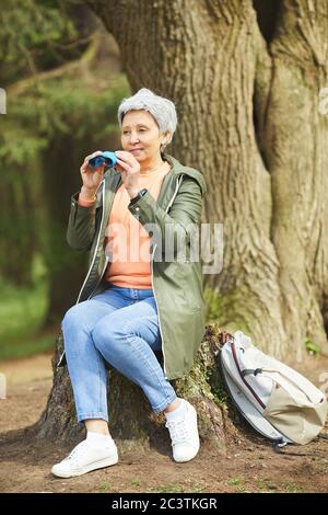 Vertikale Ganzkörperportrait von aktiven älteren Frau mit Fernglas und Lächeln während der Wanderung im Herbstwald Stockfoto