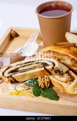 Tee in Papiertasse, Einweglöffel und Gabel, Mohn und Apfelstrudel auf einem rustikalen Holztablett Stockfoto