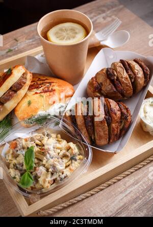 Einweggeschirr, Zitronentee, Schiefer, leckere Wurstbrötchen und Dill und gebackene Kartoffeln mit Fleisch auf einem Holztablett Seitenansicht. Stockfoto