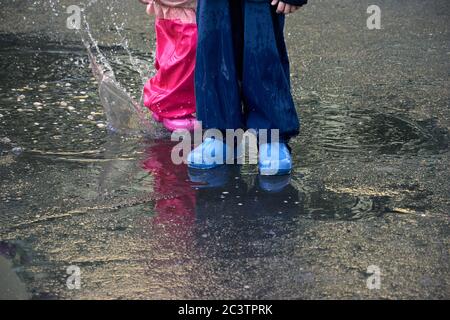 Glückliche Kinder springen in Pfütze in wasserdichtem Mantel. Junge und Mädchen haben Spaß im Regen Wetter in einem hellen Regenmantel. Stockfoto