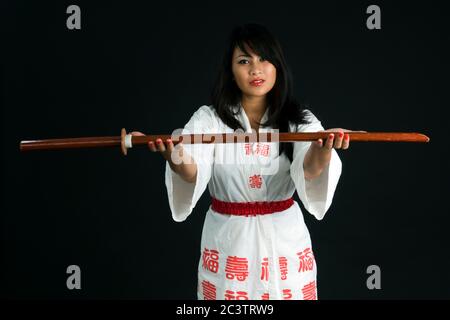 Junge asiatische Frau bietet dem Betrachter ein Bokken (ein japanisches Holzschwert, das zum Üben verwendet wird) auf schwarzem Hintergrund an Stockfoto