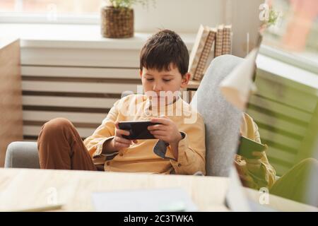 Warm getönte Porträt von niedlichen Jungen im Sessel sitzen und spielen mobile Spiele über Smartphone, kopieren Raum Stockfoto