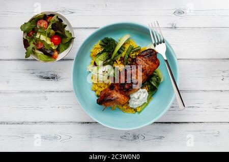 Eine köstliche Schüssel mit Tandoori-Huhn, Gemüse und Reis, in einer hellblauen Schüssel auf einem rustikalen weißen Holzhintergrund mit einem Beilagensalat geschossen Stockfoto