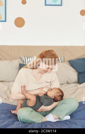 Vertikale Ganzkörperportrait von lächelnden erwachsenen Mutter Stillen gemischt Rennen Baby während auf dem Bett zu Hause sitzen, kopieren Raum Stockfoto