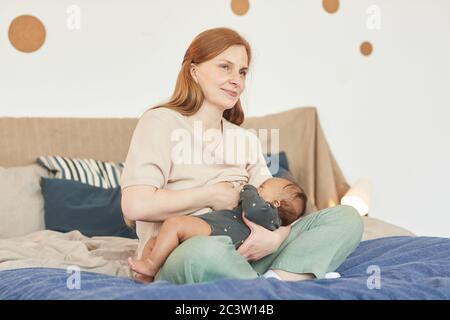 In warmen Farbtönen gehaltene Ganzkörperporträts mit lächelnder Erwachsener, die ihr Baby mit gemischtem Rennen stillt, während sie zu Hause auf dem Bett sitzen und Platz kopieren Stockfoto